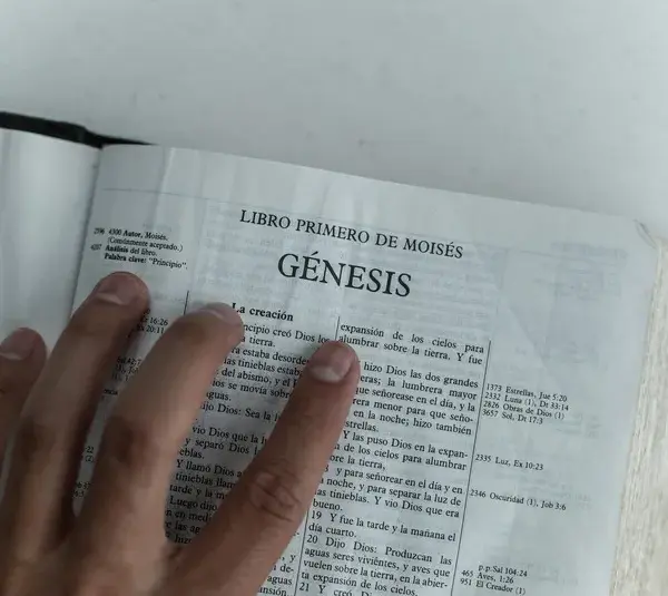 In daily devotion, a hand is placed on an open Bible on Genesis.
