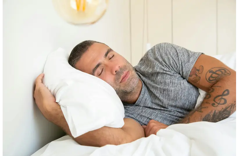 Man with tattoos peacefully sleeping on his side on a white pillow, wearing a gray t-shirt. The room exudes a calm and restful atmosphere.