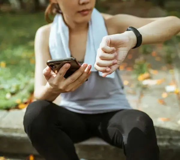 A person takes a short break outdoors, checking their fitness tracker and phone, capturing the idea of wellness stacking by combining movement, mindfulness, and health tracking in one simple moment.
