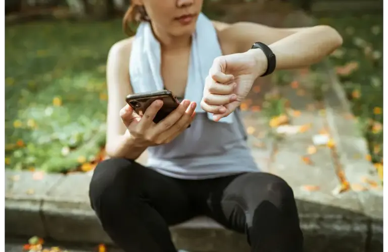 A person takes a short break outdoors, checking their fitness tracker and phone, capturing the idea of wellness stacking by combining movement, mindfulness, and health tracking in one simple moment.