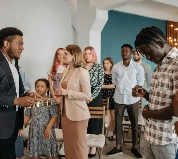 A group of people stand together in church, in a prayer or meditation setting. A man in a suit holds a collection plate. The mood is serene and focused. Group may include family and friends.