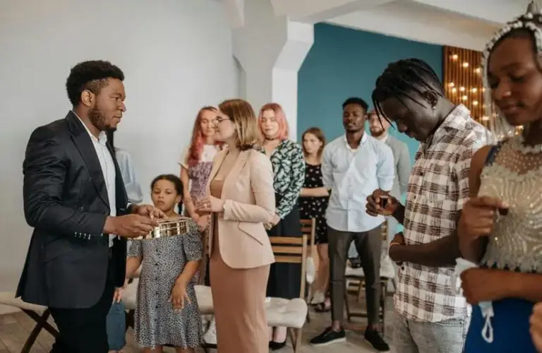 A group of people stand together in church, in a prayer or meditation setting. A man in a suit holds a collection plate. The mood is serene and focused. Group may include family and friends.