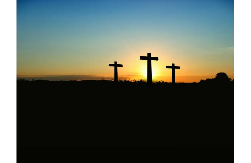 Three crosses stand silhouetted against a vibrant sunset, with a gradient sky transitioning from blue to orange, evoking a serene, contemplative mood.