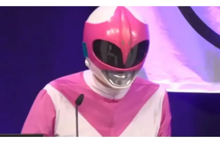 A person dressed in a pink and white superhero costume with a glossy helmet speaks at a podium. The background is a deep blue with partial lighting.