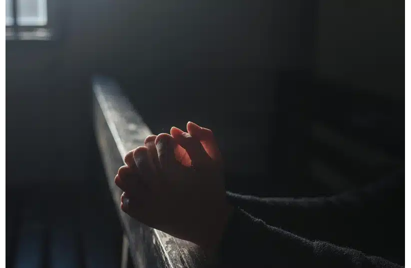 Hands clasped in prayer rest on a wooden pew, illuminated by dim sunlight from a window, conveying a serene and contemplative mood.