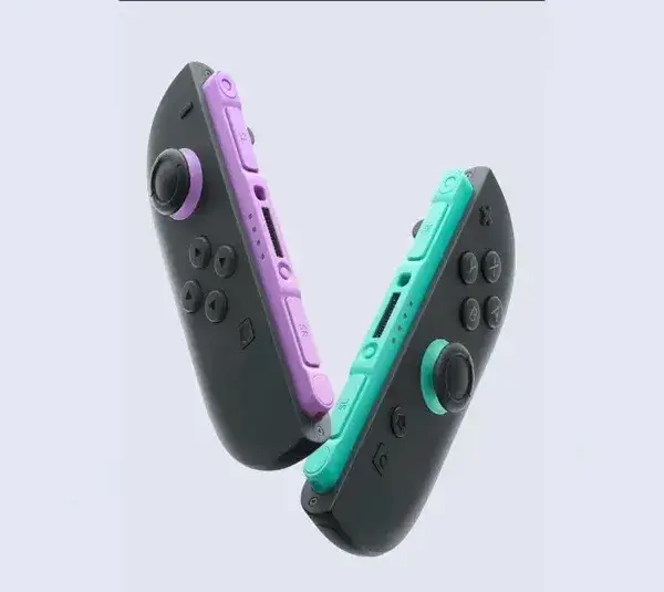 Two new Nintendo Switch 2 Joy Con controllers, one with a purple grip and the other with a teal grip, face each other against a light background, creating a playful tone.