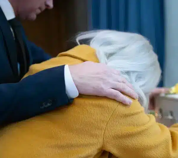 A person wearing a yellow coat leans on a coffin, visibly grieving. Another person, in a dark suit, offers comfort with a hand on their shoulder., Grief