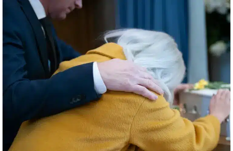 A person wearing a yellow coat leans on a coffin, visibly grieving. Another person, in a dark suit, offers comfort with a hand on their shoulder., Grief