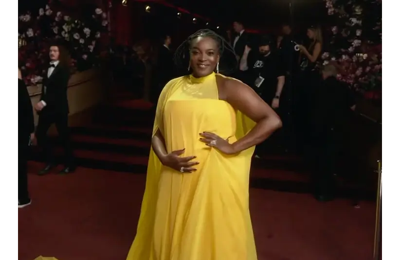Wunmi Mosaku, in a vibrant yellow cape dress, poses confidently on a red carpet. The background features elegantly dressed people and floral decorations, creating a glamorous, festive atmosphere.