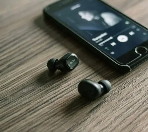 A smartphone playing music with black wireless Google fast pair earbuds beside it on a wooden surface. The scene suggests a relaxed, tech-savvy vibe.