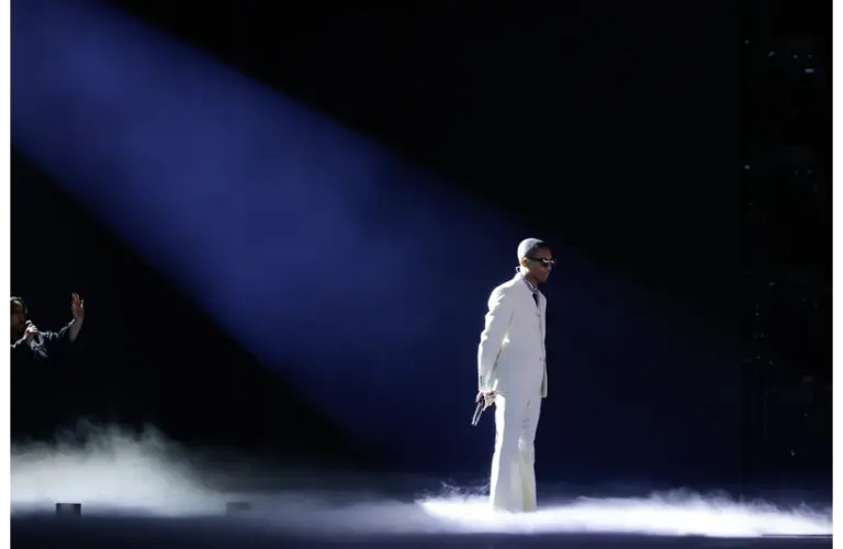 Pharrell Williams, in a white suit and sunglasses, stands in a spotlight on a smoky stage, holding a microphone. Another figure gestures in the dimly lit background.