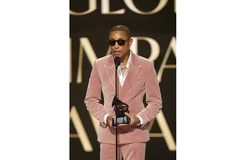 Pharrell Williams, in a pink velvet suit and sunglasses, speaks into a microphone on stage, holding an award. The background features large, out-of-focus text.