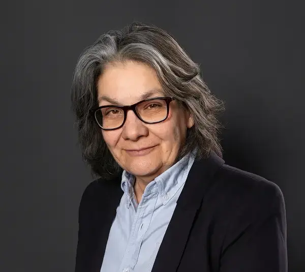 Ann Godoff, legendary editor, a person with gray hair and glasses smiles slightly. They wear a light blue shirt and dark blazer, set against a plain dark background, conveying warmth and professionalism.