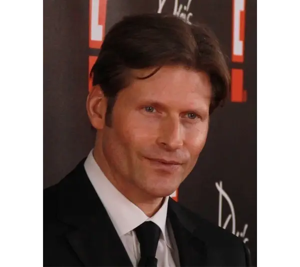An image of Crispin Glover in a black suit and tie is smiling slightly against a dark backdrop with red and white patterns, creating a formal and composed mood.
