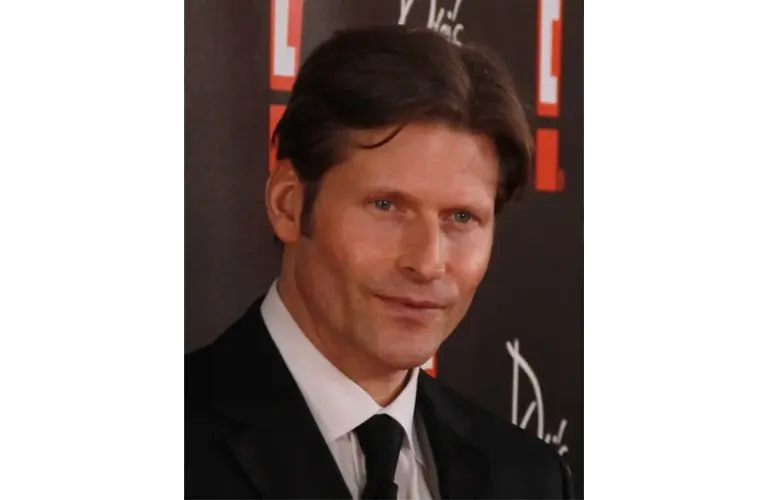 An image of Crispin Glover in a black suit and tie is smiling slightly against a dark backdrop with red and white patterns, creating a formal and composed mood.