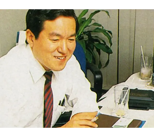 Hideki Sato, SEGA engineer, in a white shirt and red tie smiles while sitting at a desk. He appears to be focused on his work. A television and a plant are in the background.