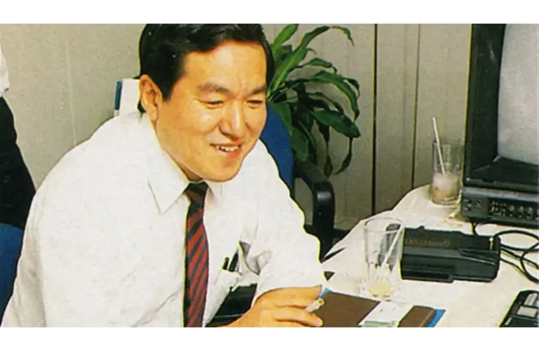 Hideki Sato, SEGA engineer, in a white shirt and red tie smiles while sitting at a desk. He appears to be focused on his work. A television and a plant are in the background.
