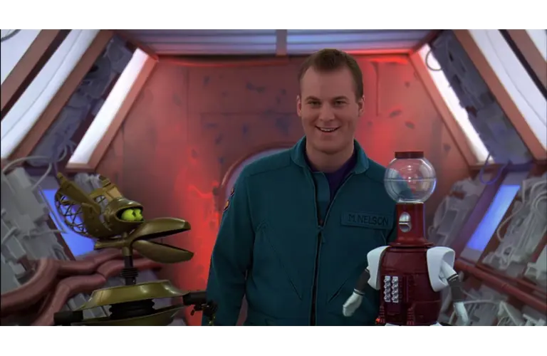 Mike Nelson, in a teal jumpsuit, smiles warmly between two robots, Tom Servo (on the right) and Crow T. Robot (on the left), on the Satellite of Love spaceship setting for MST3K. The mood is playful and light-hearted.