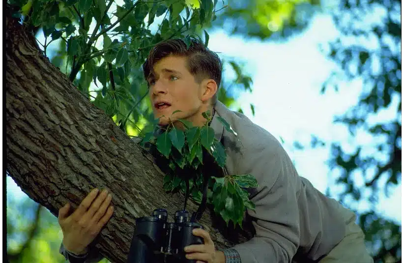 Crispin Glover, as a young man clutches a tree branch with binoculars in hand, looking anxiously to the side. Sunlight filters through leaves, creating a tense, watchful mood.