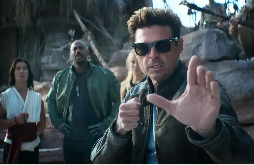 A man in sunglasses and a leather jacket gestures energetically at the camera, surrounded by three others in varied costumes, set against a rocky backdrop.