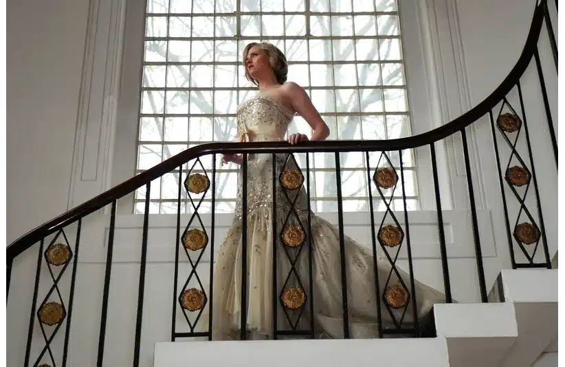 Kristen Stewart as Diana, Princess of Wales, in an elegant, bejeweled gown, stands on a curved staircase, bathed in light from a large window. The scene conveys elegance and poise.
