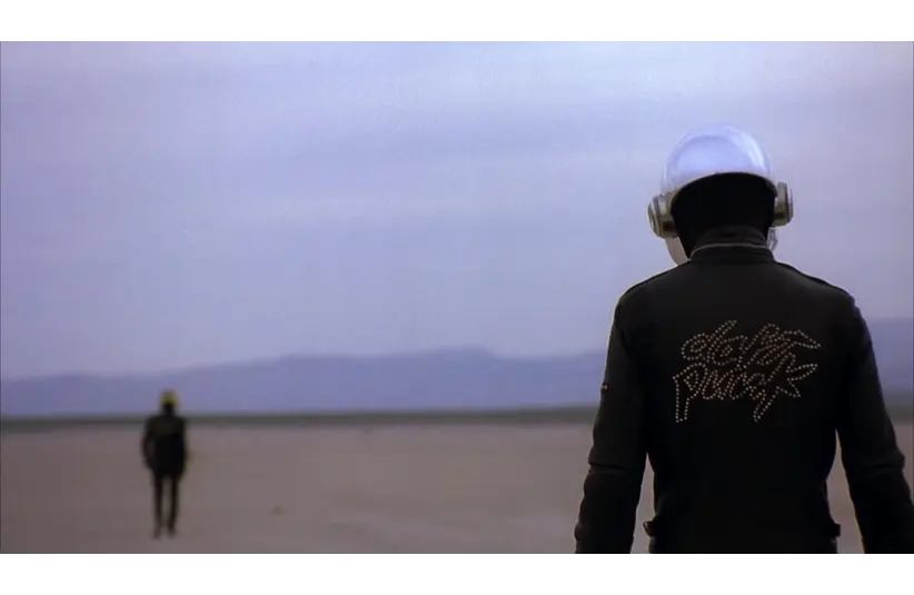 A futuristic figure in a black jacket with "Daft Punk" on the back faces an expansive desert landscape; another figure is in the distance, under a cloudy sky.