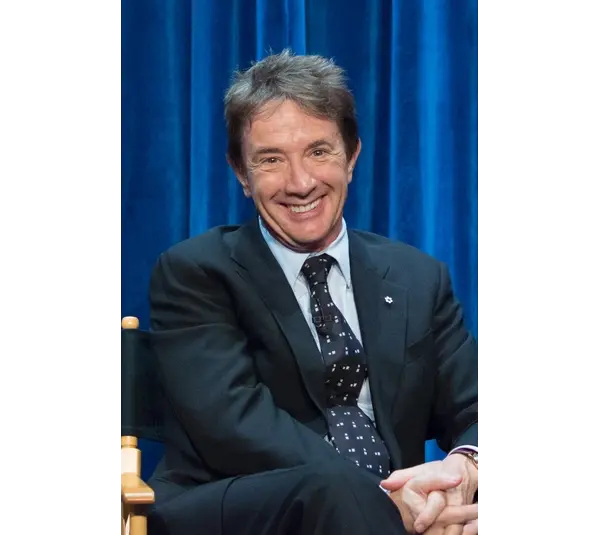 Martin Short, father of the late Katherine H. Short, sits in a suit sits smiling against a vibrant blue curtain backdrop. Their relaxed posture and cheerful expression convey a friendly, welcoming tone. The Short family is in mourning.