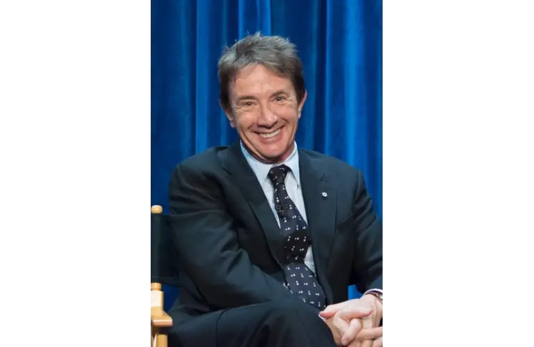 Martin Short, father of the late Katherine H. Short, sits in a suit sits smiling against a vibrant blue curtain backdrop. Their relaxed posture and cheerful expression convey a friendly, welcoming tone. The Short family is in mourning.