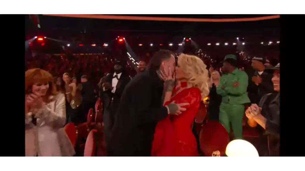 Jelly Roll and wife, Bunnie Xo, share a kiss in a crowded theater, surrounded by applauding people. The woman is in a red dress and the man in a black jacket. The atmosphere is joyful and celebratory.