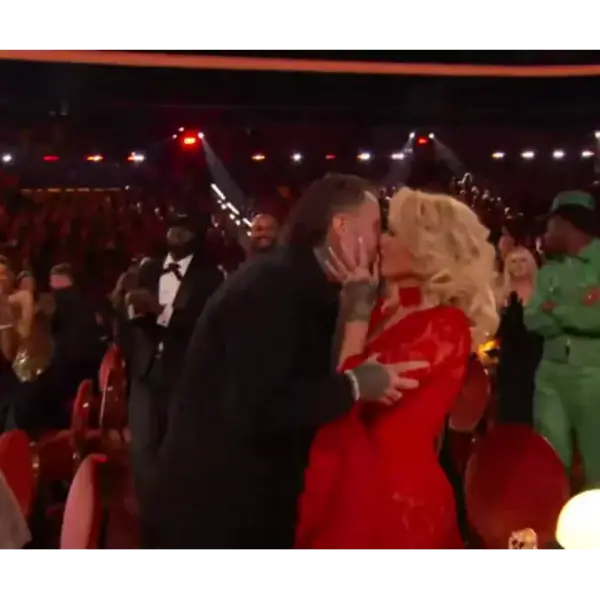 Jelly Roll and wife, Bunnie Xo, share a kiss in a crowded theater, surrounded by applauding people. The woman is in a red dress and the man in a black jacket. The atmosphere is joyful and celebratory.