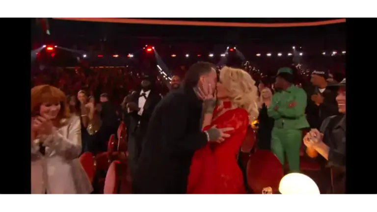 Jelly Roll and wife, Bunnie Xo, share a kiss in a crowded theater, surrounded by applauding people. The woman is in a red dress and the man in a black jacket. The atmosphere is joyful and celebratory.