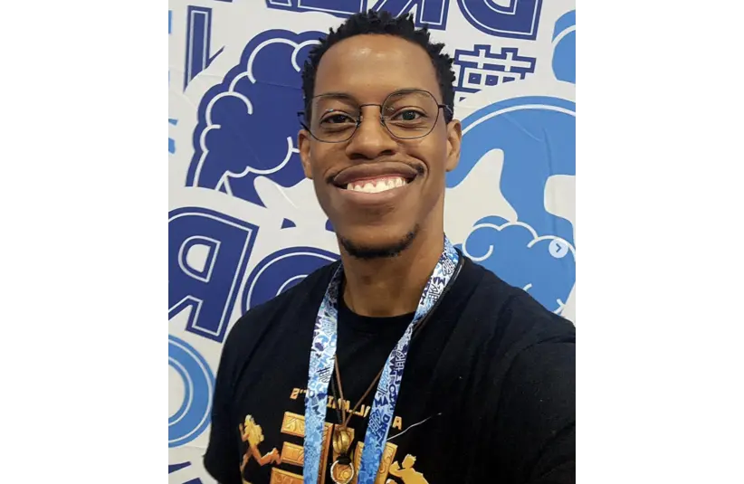 Aerial Knight smiles broadly while standing indoors. He wears glasses and a black shirt with a colorful design. A blue and white patterned lanyard hangs around his neck. The background features cartoonish graphics in blue and white, creating a lively, upbeat atmosphere.