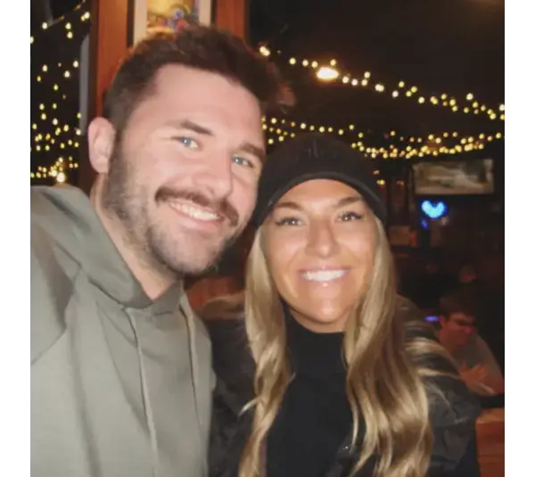 Screenshot of Brianna “Bri” McNees and Connor Spies, Courtesy of breezy_mcneezy via Instagram, shows a smiling man and woman pose for a selfie in a dimly lit room with string lights in the background, creating a warm and cheerful atmosphere.