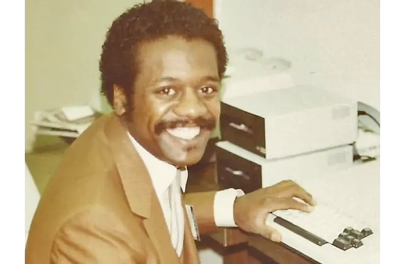 Edward Smith, creator of the first pc-like gaming system, the imagination machine is in a brown suit sits at a vintage computer, typing. The scene conveys a positive, professional atmosphere in an office setting.