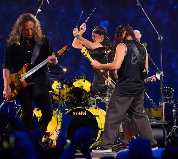 Metallica, a rock band performs energetically on stage. One guitarist is strumming intensely, another faces the drummer, who is mid-action. Their backdrop is a vast, cheering crowd.