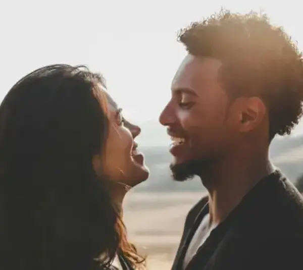 A couple stands closely, smiling at each other against a sunlit, blurred countryside. The mood is warm and joyful, capturing an intimate, happy moment, emotional clarity