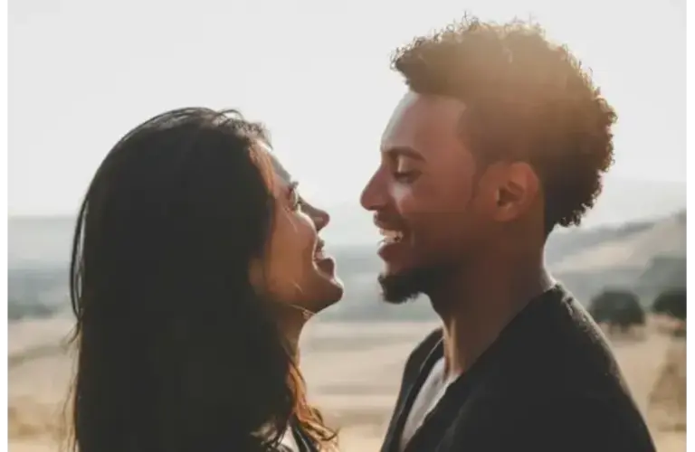 A couple stands closely, smiling at each other against a sunlit, blurred countryside. The mood is warm and joyful, capturing an intimate, happy moment, emotional clarity