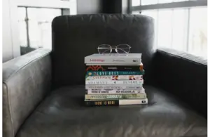 A stack of books with glasses rests on a dark gray armchair. Sunlight filters through a window, casting soft shadows. The scene conveys a cozy, intellectual atmosphere with best sellers to choose from.