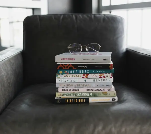 A stack of books with glasses rests on a dark gray armchair. Sunlight filters through a window, casting soft shadows. The scene conveys a cozy, intellectual atmosphere with best sellers to choose from.