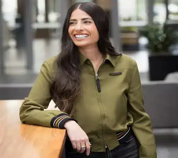 Smiling woman, Asha Sharma, with long dark hair leans against a wooden table, wearing an olive green jacket. Office setting with a relaxed, cheerful atmosphere.