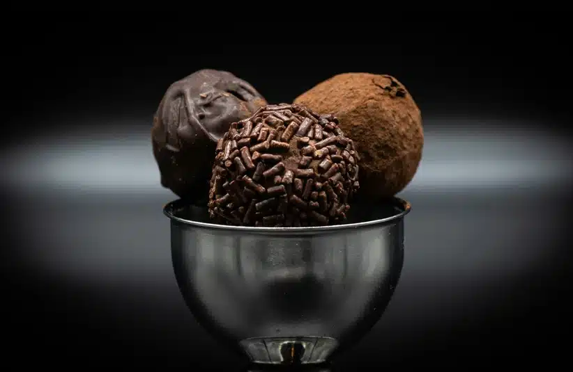 A trio of chocolate truffles sits in a silver cup against a dark background. Each truffle has a distinct texture: smooth, coated with sprinkles, and dusted with cocoa.