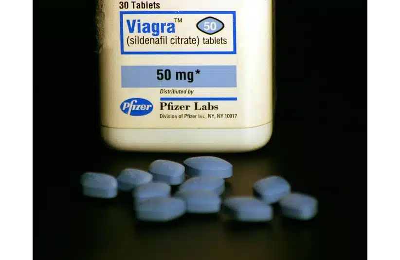 A bottle of Viagra tablets is placed on a table with several blue pills scattered in front. The label reads "50 mg," showing a medical context.
