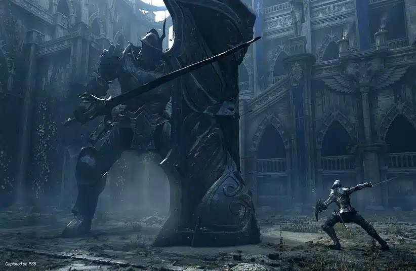 A knight in silver armor, in Bluepoint Games Remake game Demon's Souls battles a towering armored giant wielding a massive sword in a gothic, dilapidated cathedral. The scene evokes tension.