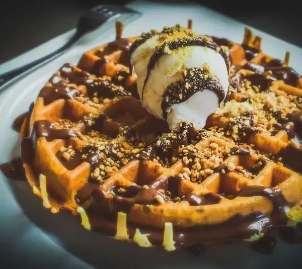 image of A warm waffle drizzled with chocolate sauce, topped with a scoop of vanilla ice cream and sprinkled with crushed nuts, sits on a white plate. Indulgent and inviting for Galentine's Day brunch