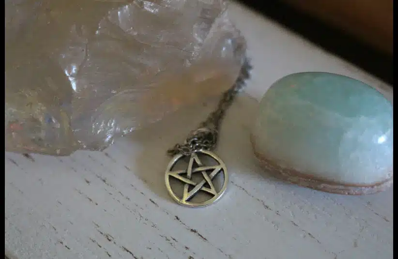 A silver pentacle necklace lies between a large translucent crystal and a smooth, light blue stone on a rustic white wooden surface, conveying a mystical and serene atmosphere. Wiccan Rede