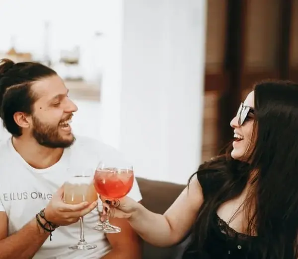 image of A man and woman smile and clink glasses filled with colorful drinks, conveying joy and celebration in a relaxed, indoor setting for Valentine's Day