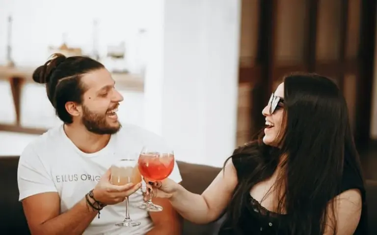 image of A man and woman smile and clink glasses filled with colorful drinks, conveying joy and celebration in a relaxed, indoor setting for Valentine's Day