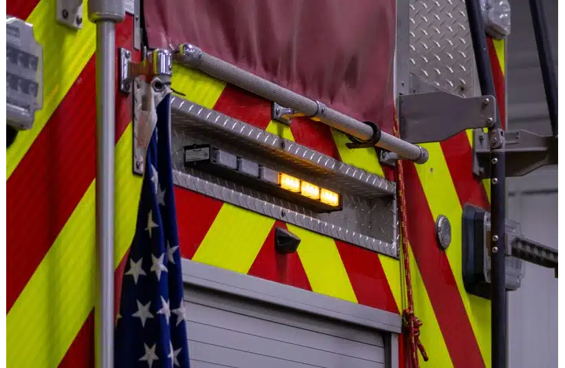 Close-up of a fire truck's rear, showing red and yellow chevron pattern, reflective striping, an American flag, and a lit emergency light.