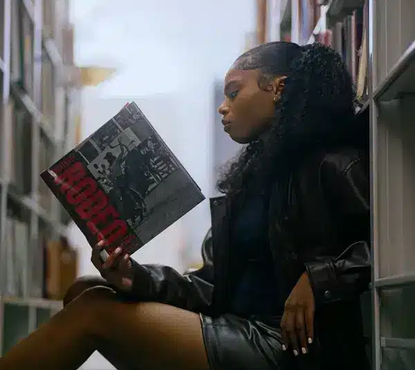 Young woman in a leather jacket sits on a library floor, reading a magazine titled "Rodeo" for Black History Month. She appears focused, surrounded by tall shelves of books.