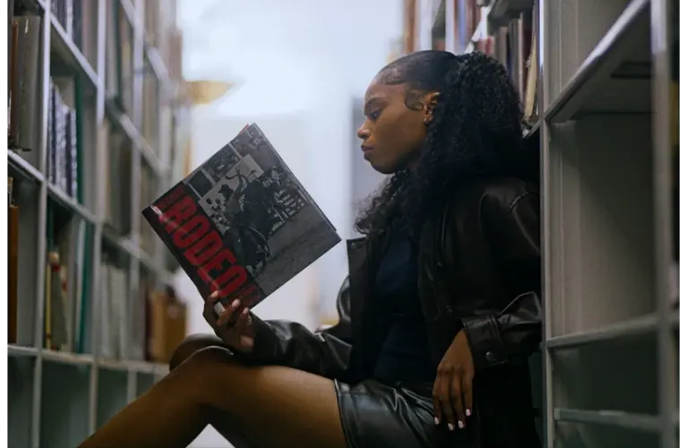 Young woman in a leather jacket sits on a library floor, reading a magazine titled "Rodeo" for Black History Month. She appears focused, surrounded by tall shelves of books.
